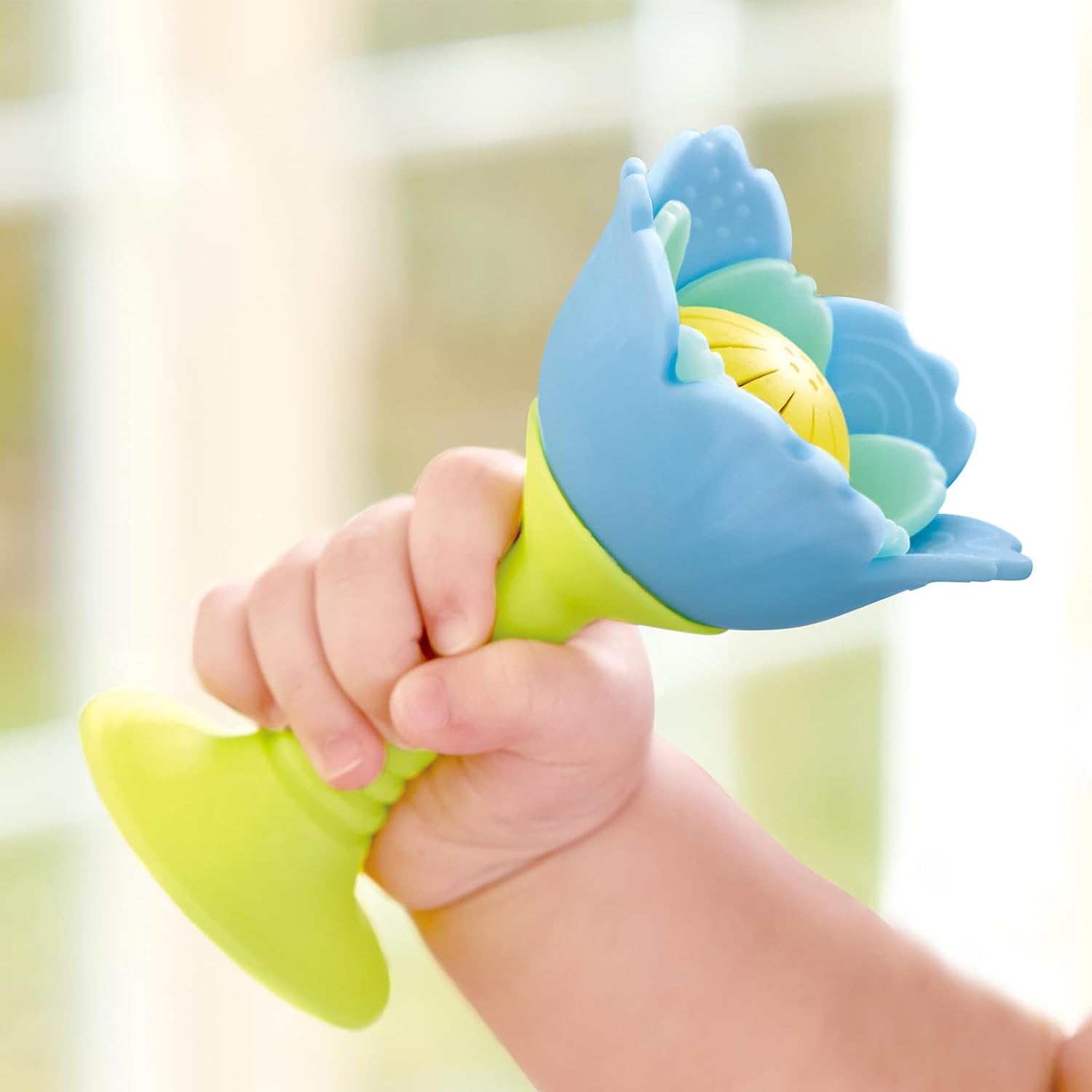 A baby’s hand holds the Hape Rattle & Teether Blue, a silicone flower toy with chewy petals and a yellow ball center—perfect for sensory play—set against a softly blurred background.