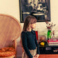 A young girl in a dark top and Louis Louise's Girls' Zippa Corduroy Leopard Skirt stands indoors by a wicker chair and sideboard decorated with books, flowers, and a large framed photo.