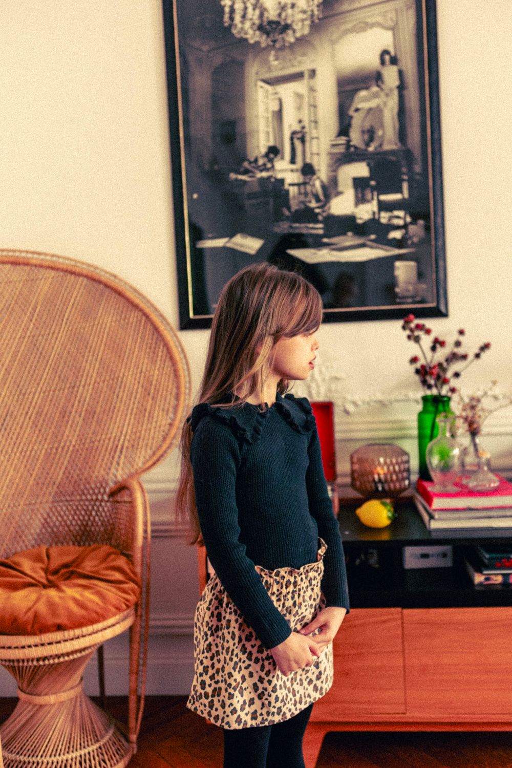 A young girl in a dark top and Louis Louise's Girls' Zippa Corduroy Leopard Skirt stands indoors by a wicker chair and sideboard decorated with books, flowers, and a large framed photo.