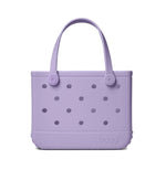 The Bitty Bogg® Bag by Bogg Bags is a compact lavender tote with round holes and two handles, featuring “bogg” embossed on the bottom right corner. This washable bag is perfect for everyday use.