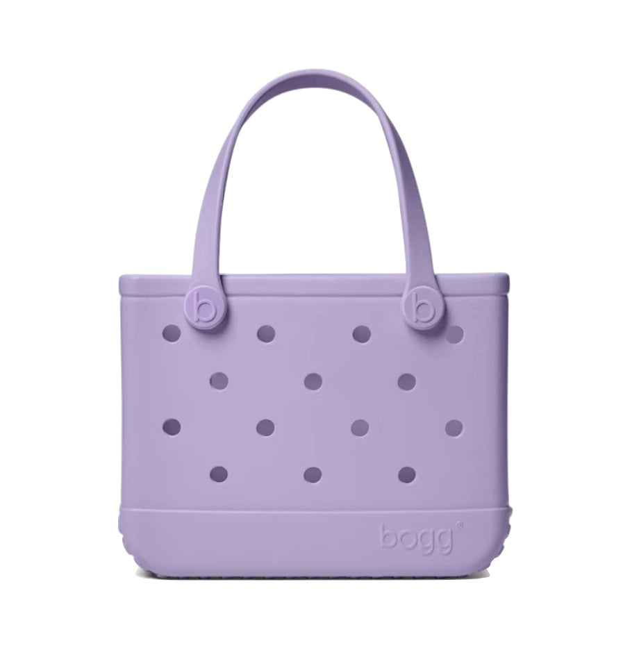 The Bitty Bogg® Bag by Bogg Bags is a compact lavender tote with round holes and two handles, featuring “bogg” embossed on the bottom right corner. This washable bag is perfect for everyday use.