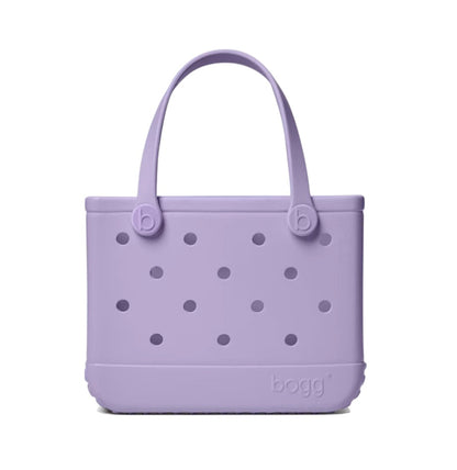 The Bitty Bogg® Bag by Bogg Bags is a compact lavender tote with round holes and two handles, featuring “bogg” embossed on the bottom right corner. This washable bag is perfect for everyday use.