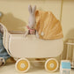 A Maileg My Rabbit, Green sitting in a miniature white pram with a yellow checkered cover next to a toy bear and a blue block spelling "milk.