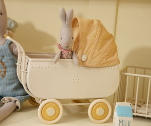 A Maileg My Rabbit, Green sitting in a miniature white pram with a yellow checkered cover next to a toy bear and a blue block spelling "milk.