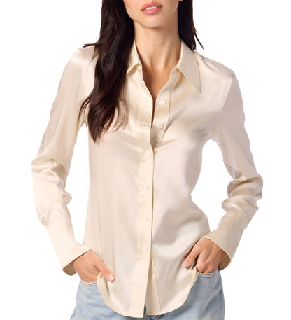 A person is wearing a long-sleeve, cream-colored satin blouse with their hands in their jean pockets, reminiscent of the elegant simplicity of the Sablyn Vincent Silk Shirt by Sablyn.