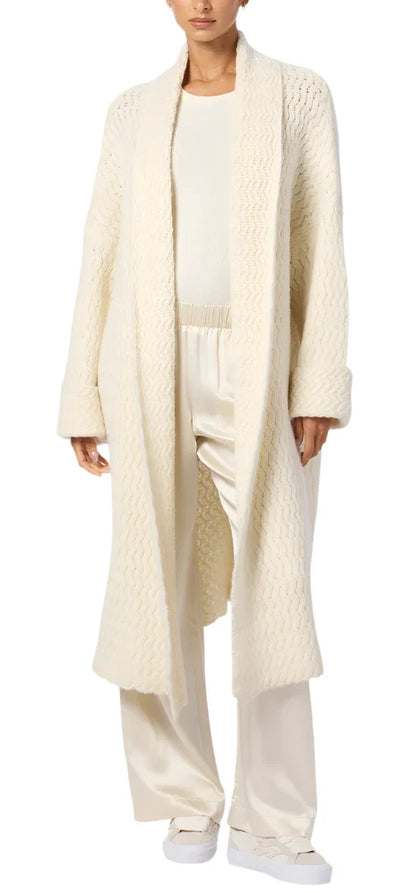 A person wearing a long, cream-colored Sablyn Stanton Slouchy Duster Cardigan with a wavy knit texture over a matching top and silk pants, complemented by slouchy design white shoes.