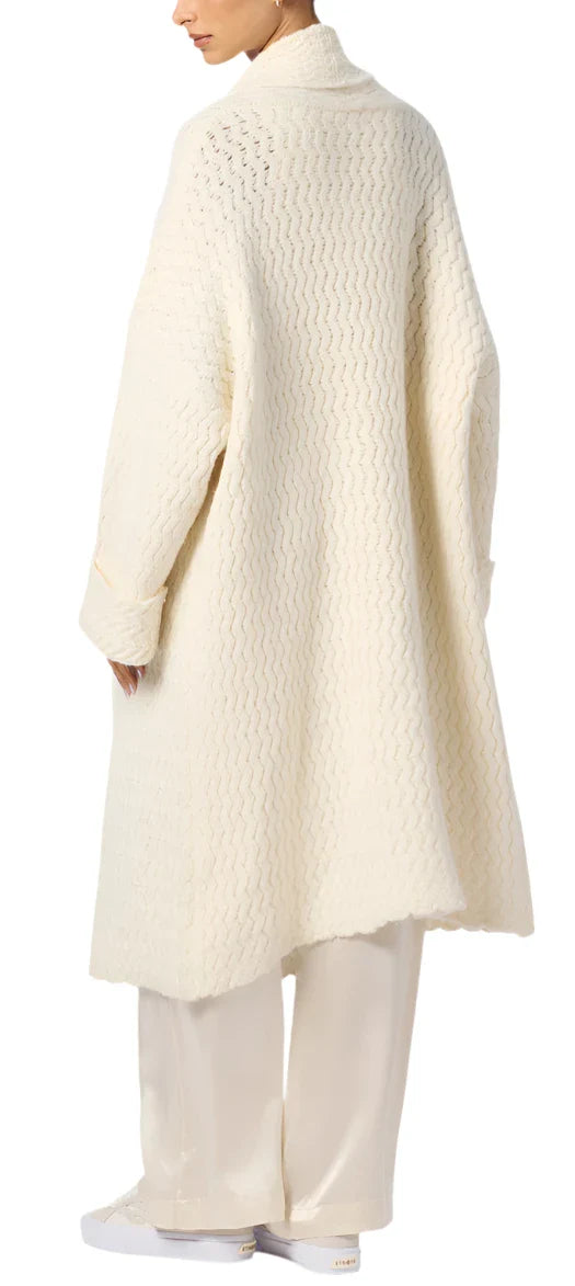 A person is seen from the back, wearing a long, textured white coat with a wavy knit pattern over white pants, reminiscent of the Sablyn Stanton Slouchy Duster Cardigan.