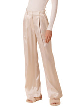 A person wearing the Sablyn Emerson Pleated Silk Pant, featuring high-rise and wide-leg design in beige, paired with fluffy slippers, posing against a white background.