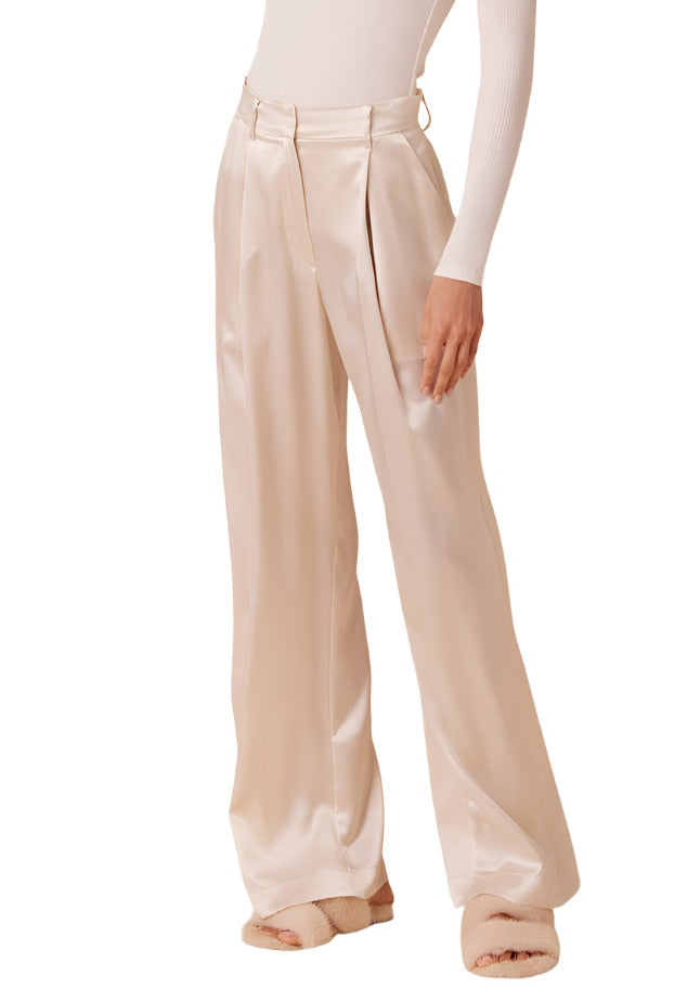 A person wearing the Sablyn Emerson Pleated Silk Pant, featuring high-rise and wide-leg design in beige, paired with fluffy slippers, posing against a white background.