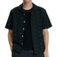 A person wears the Avery Embroidered Camp Shirt by Rag & Bone in black cotton-linen over a black ribbed t-shirt, standing with hands in pockets against a plain background.