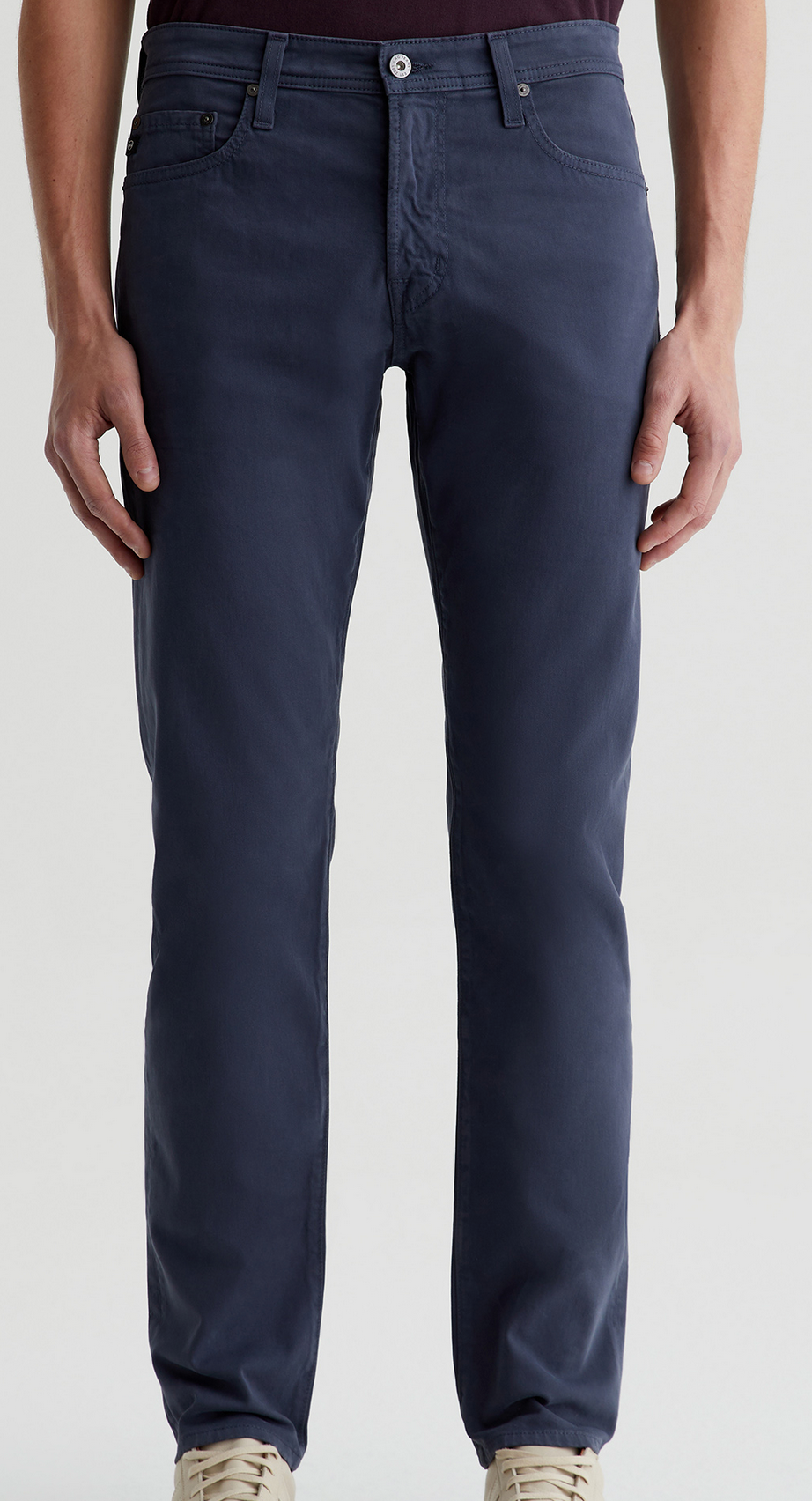 A person wearing AG Jeans Tellis Sueded Sateen slim pants and beige sneakers stands against a plain light background.