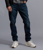 A person wearing Rag & Bone’s Fit 3 Authentic Stretch dark blue denim jeans with a tapered leg, a striped shirt, a black jacket, and white sneakers stands against a plain gray background.