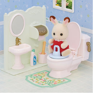 A Calico Critters Toilet Seat is featured in a miniature bathroom scene with a toy rabbit in a red dress, surrounded by a sink, mirror, cleaning accessories, and a patterned rug.