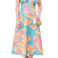 A woman in a colorful floral dress, known as the Lela Rose Isabel Dress, stands while holding a small handbag. She wears sandals and has long hair partly styled over one shoulder. The painterly print of the silky V-neck dress adds a touch of elegance to her look.