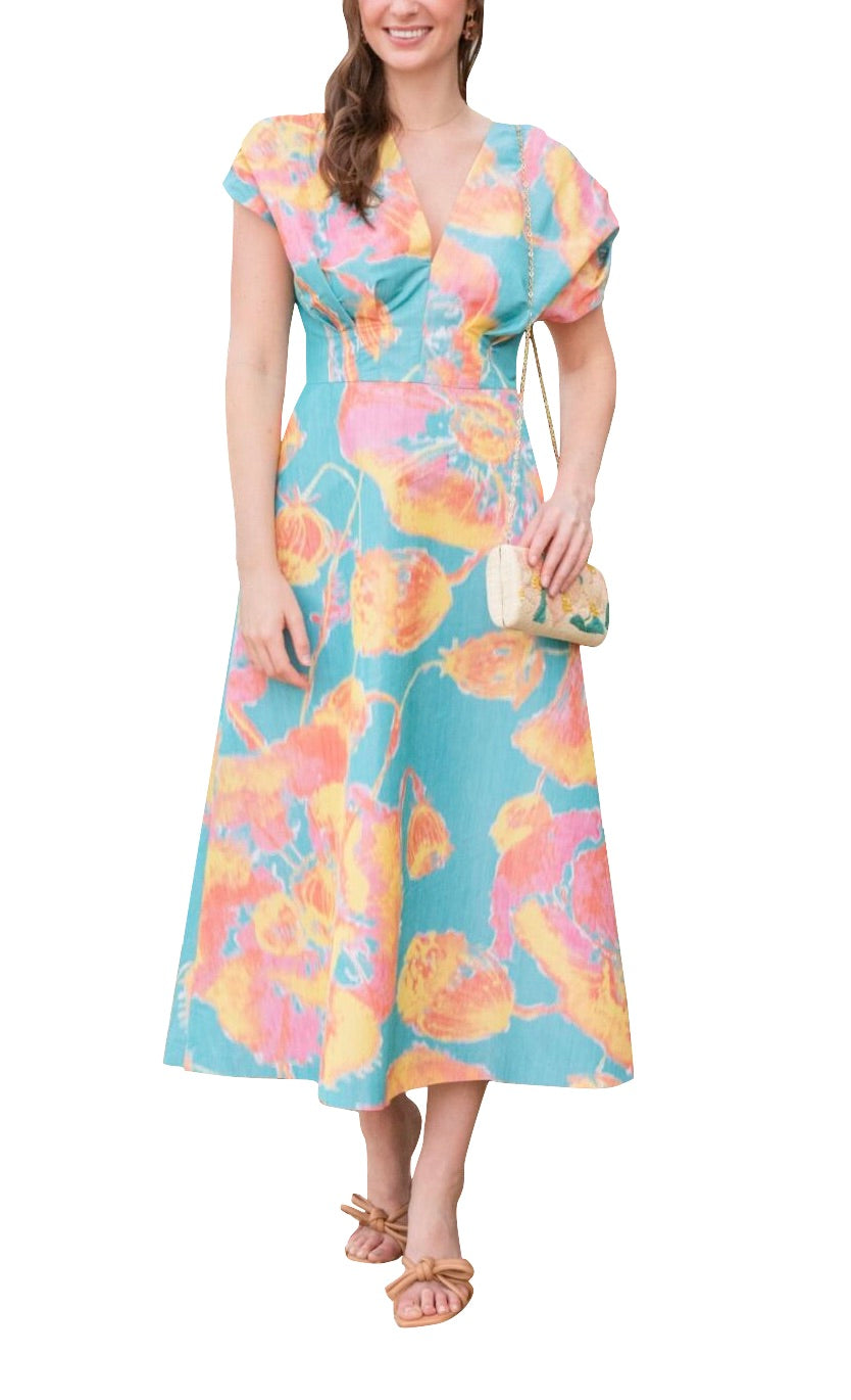 A woman in a colorful floral dress, known as the Lela Rose Isabel Dress, stands while holding a small handbag. She wears sandals and has long hair partly styled over one shoulder. The painterly print of the silky V-neck dress adds a touch of elegance to her look.