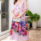 A woman wearing a flowy Lela Rose Watercolor Floral Cotton Voile Isabel Dress and holding a light blue clutch stands on a gravel path near a sliding glass door with potted plants in the background.