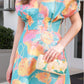 A woman is wearing a colorful floral **Lela Rose Isabel Dress** with a painterly print and holding a small, woven handbag with an embroidered design. She is standing on a patio near a doorway.