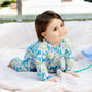 A toddler in Magnetic Me's "Baby' It's a Paw-ty" Convertible Coverall, featuring an animal pattern and flame-resistant fabric, crawls on a white outdoor blanket, glancing to the side with a slight smile.