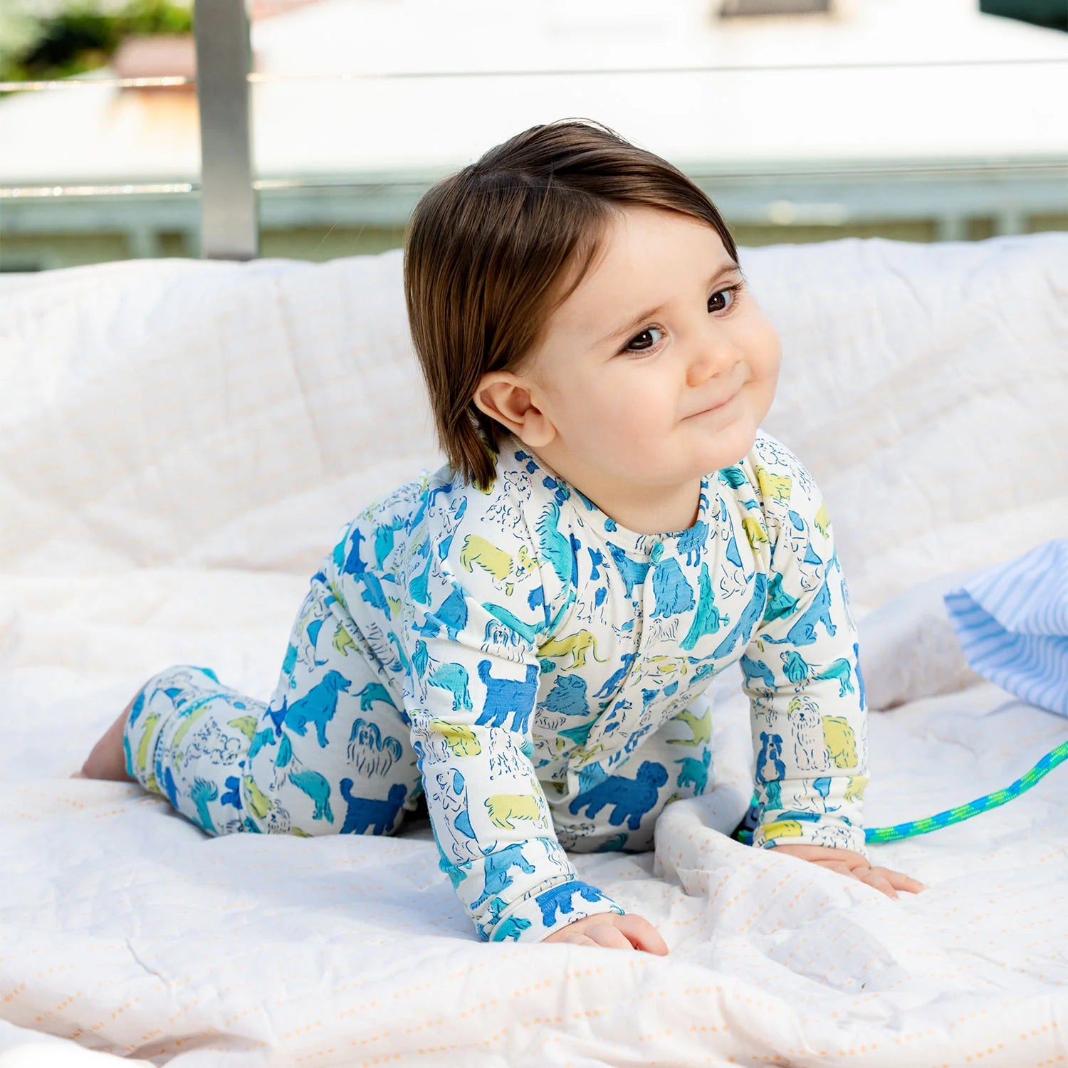 A toddler in Magnetic Me's "Baby' It's a Paw-ty" Convertible Coverall, featuring an animal pattern and flame-resistant fabric, crawls on a white outdoor blanket, glancing to the side with a slight smile.