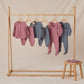 A wooden rack displays five Quincy Mae Baby Boys' and Boys' Sweatpants in mauve and blue tones with elastic waistbands; a stool with folded clothes stands to the right on a beige backdrop.