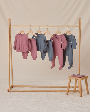 A wooden rack displays five Quincy Mae Baby Boys' and Boys' Sweatpants in mauve and blue tones with elastic waistbands; a stool with folded clothes stands to the right on a beige backdrop.