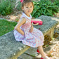 A young girl in a Kidiwi Baby Girls' and Girls' Ixia Smocked Dress and red shoes sits on a stone bench outdoors, wearing a feathered headband and looking slightly to the side.