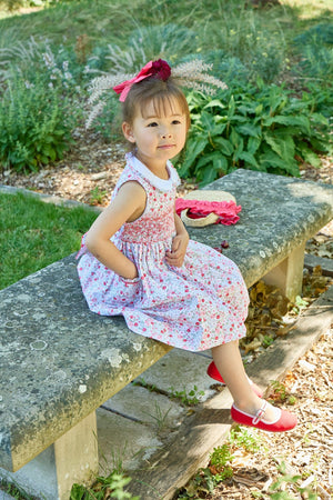 A young girl in a Kidiwi Baby Girls' and Girls' Ixia Smocked Dress and red shoes sits on a stone bench outdoors, wearing a feathered headband and looking slightly to the side.