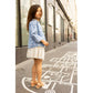 A young girl wearing the Louis Louise Girls’ Brune Denim Flower Jacket stands on a sidewalk with a hopscotch game drawn on it, glancing slightly over her shoulder.