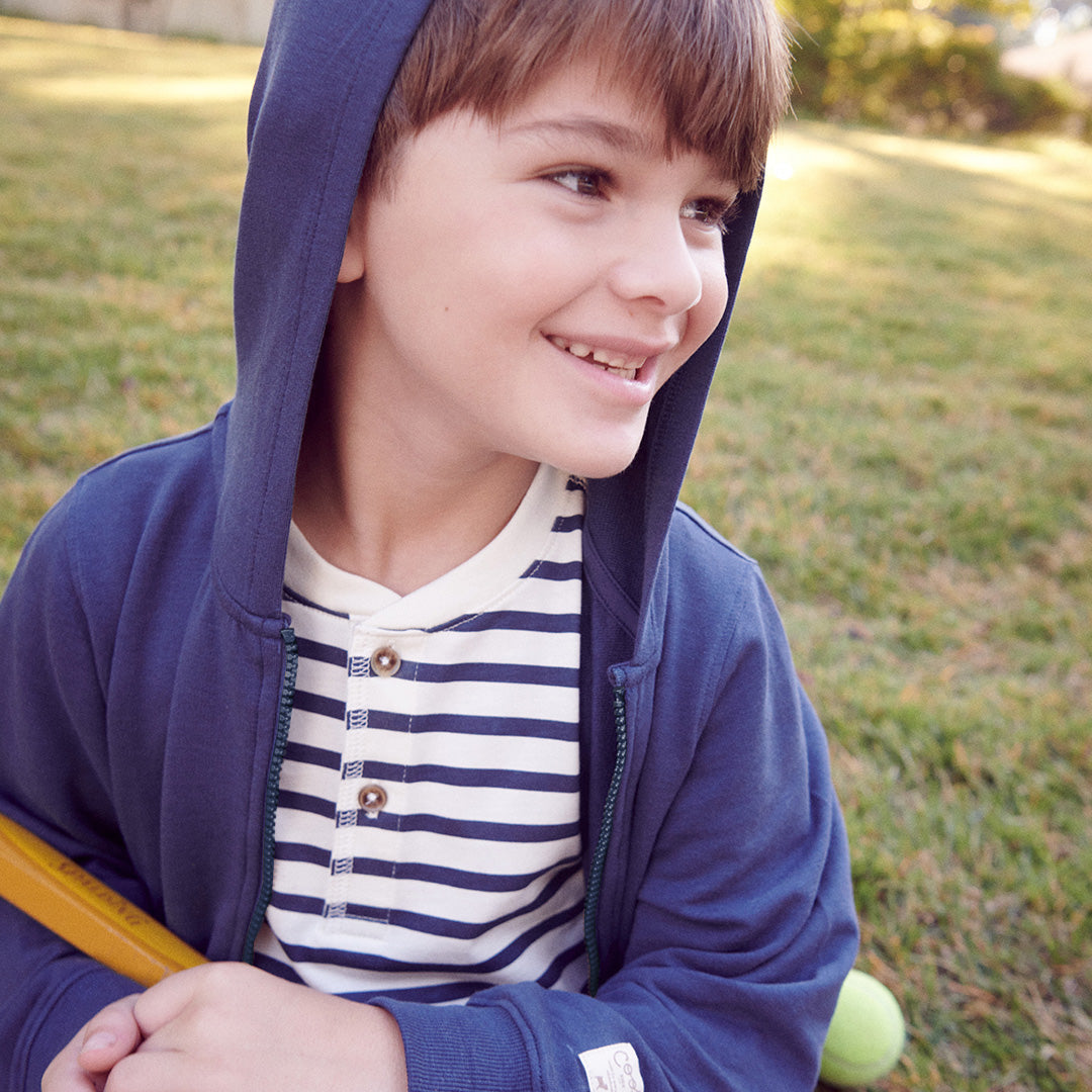 A smiling child wears the Coop Kids PC Kids’ Jax Hoodie in blue over a striped shirt, sitting on the grass holding a yellow bat, with a green ball nearby.