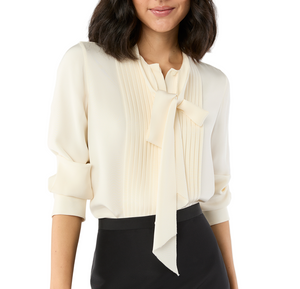 A woman wears the Ann Mashburn Marigold Blouse in cream crepe de chine, featuring pleat detailing and a bow tie, styled with a black skirt.