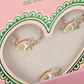 The Konges Slojd Jewelery Set comes in a pink box with a heart-shaped window, featuring gold stars and rainbow designs framed by a green scalloped edge.