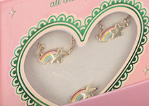 The Konges Slojd Jewelery Set comes in a pink box with a heart-shaped window, featuring gold stars and rainbow designs framed by a green scalloped edge.