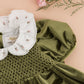 The Kidiwi Baby Girls’ and Girls’ Joséphine Smocked Dress in olive green features puff sleeves and an embroidered white ruffled collar with pink floral details, displayed on a beige surface with pink carnations above.