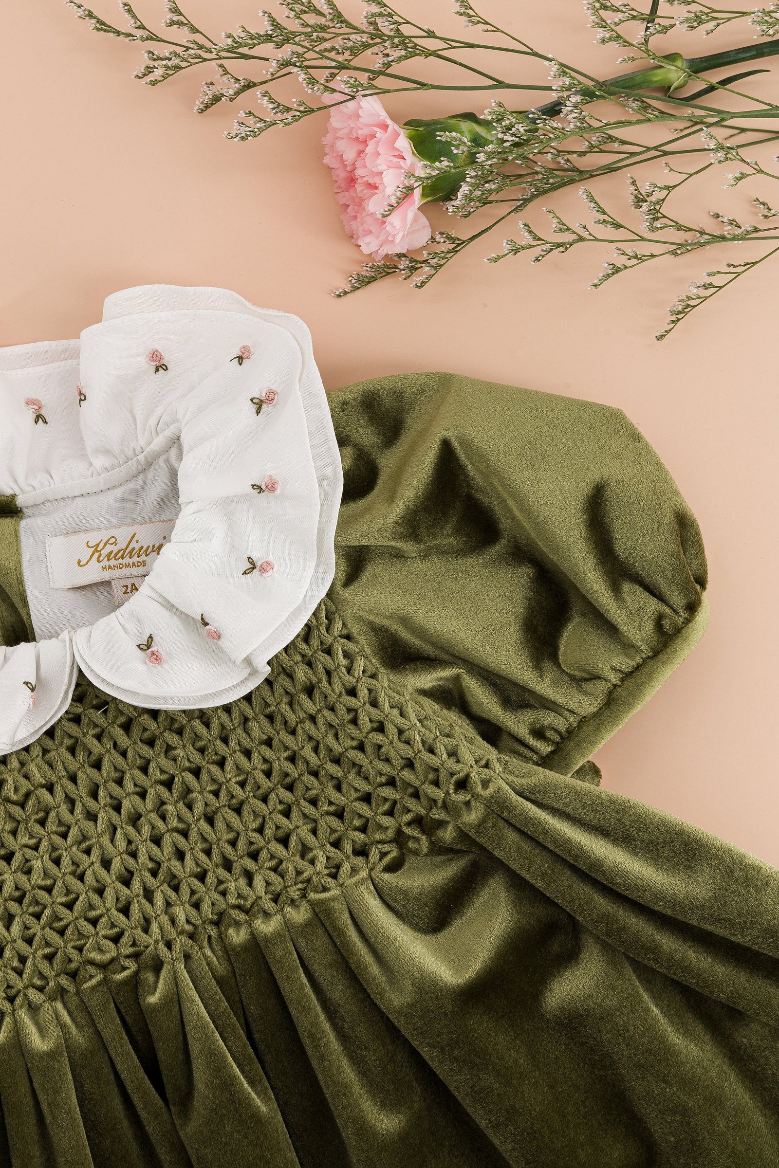 The Kidiwi Baby Girls’ and Girls’ Joséphine Smocked Dress in olive green features puff sleeves and an embroidered white ruffled collar with pink floral details, displayed on a beige surface with pink carnations above.