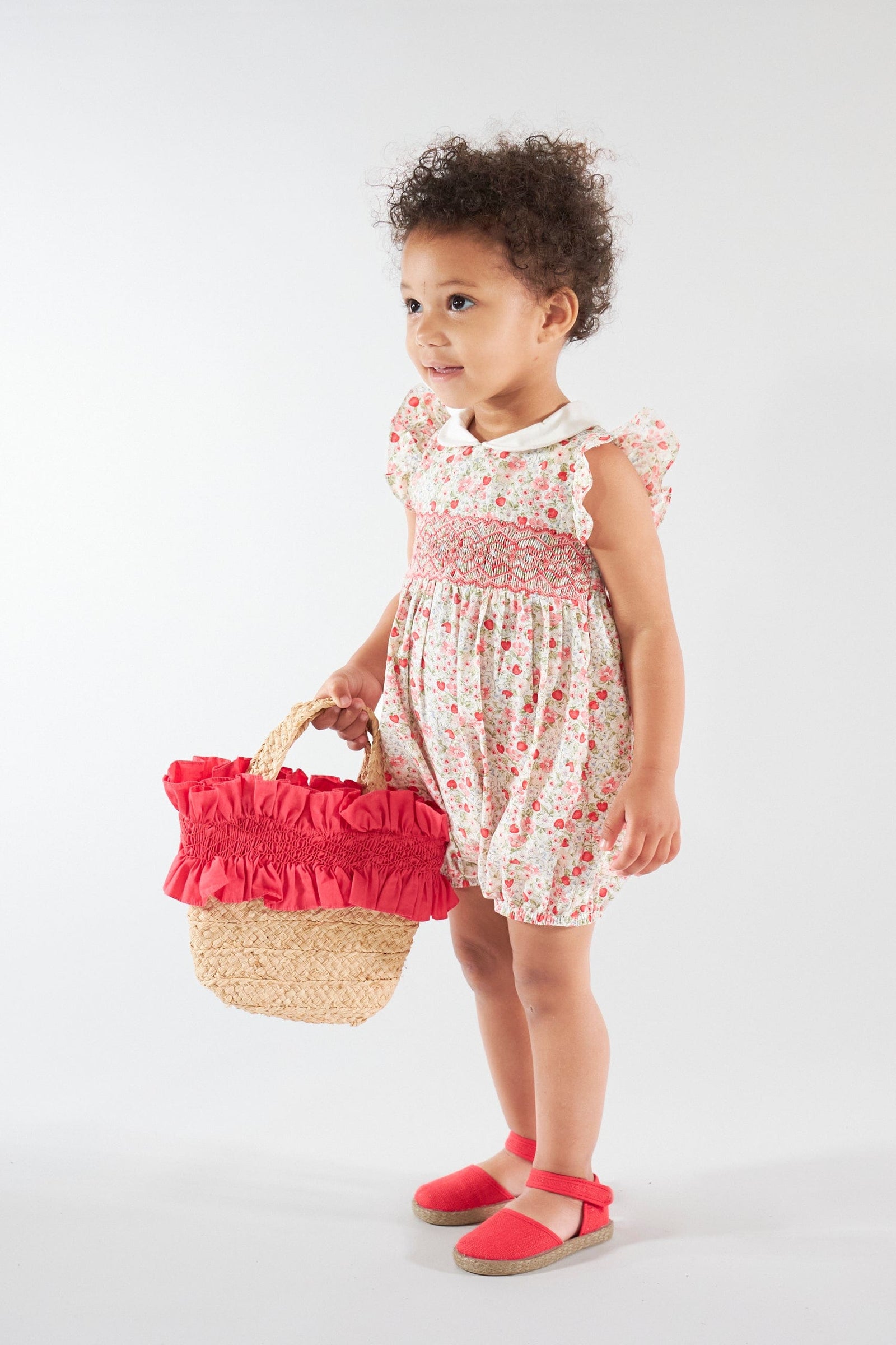 A young child wears the Kidiwi Baby Girls' Julyne Smocked Girl Romper and red shoes, holding a straw basket with red fabric trim, against a plain white background.