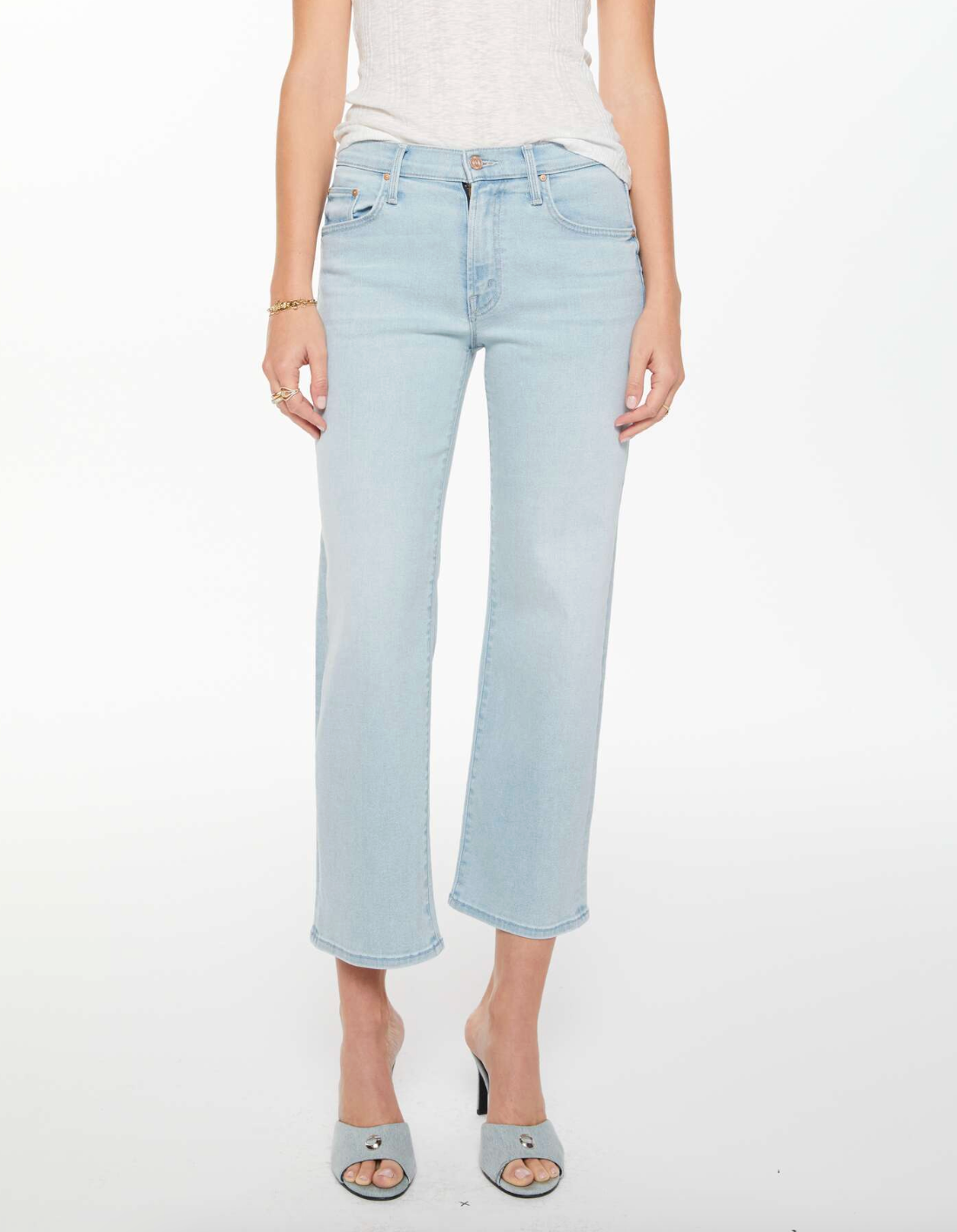 A person wearing Mother’s Mid Rise Rambler Zip Ankle Fray jeans, a white sleeveless top, and open-toe heeled sandals against a white background.