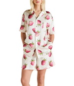 A woman stands against a white background, hands in pockets, wearing the Olivia von Halle Emeli Silk Pajamas—a white short-sleeved set adorned with a playful pink strawberry print.