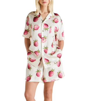 A woman stands against a white background, hands in pockets, wearing the Olivia von Halle Emeli Silk Pajamas—a white short-sleeved set adorned with a playful pink strawberry print.