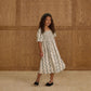 A young girl with long curly hair stands on a beige carpet in front of a wooden panel wall, wearing the Rylee & Cru Girls' Della Dress with a smocked bodice and black shoes.