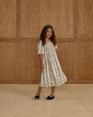 A young girl with long curly hair stands on a beige carpet in front of a wooden panel wall, wearing the Rylee & Cru Girls' Della Dress with a smocked bodice and black shoes.