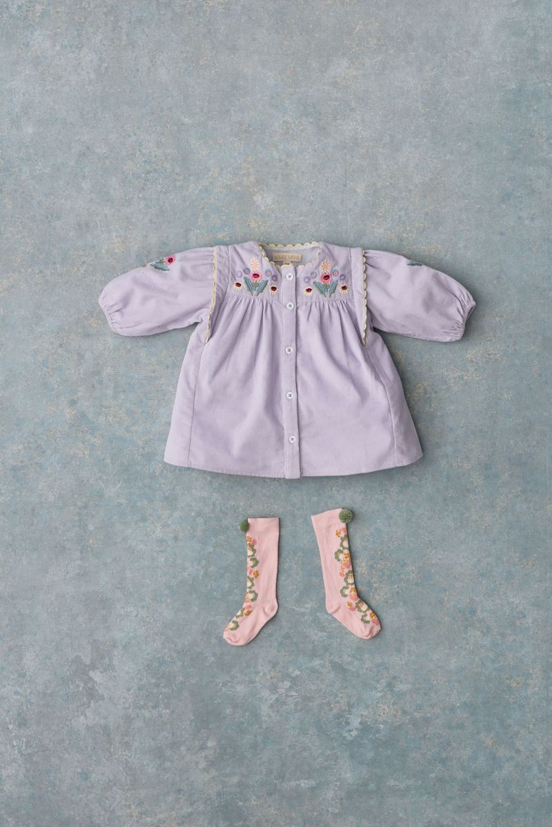 The Louise Misha Baby Girls’ and Girls’ Anette Dress is shown here in lavender corduroy with an embroidered yoke and subtle ric rac details, paired with pink knee-high socks. Both items are laid flat on a textured light blue background.
