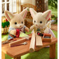 Two Fennec Fox Family figures from Calico Critters, dressed in fabric clothing, use toy tools and wood pieces on a small table outdoors, surrounded by grass and garden-themed accessories.