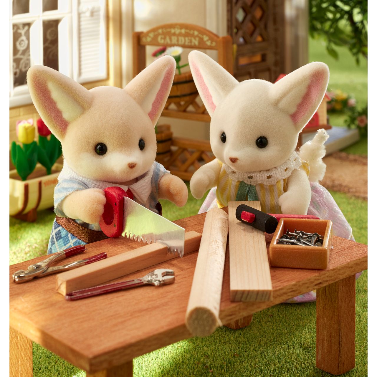 Two Fennec Fox Family figures from Calico Critters, dressed in fabric clothing, use toy tools and wood pieces on a small table outdoors, surrounded by grass and garden-themed accessories.