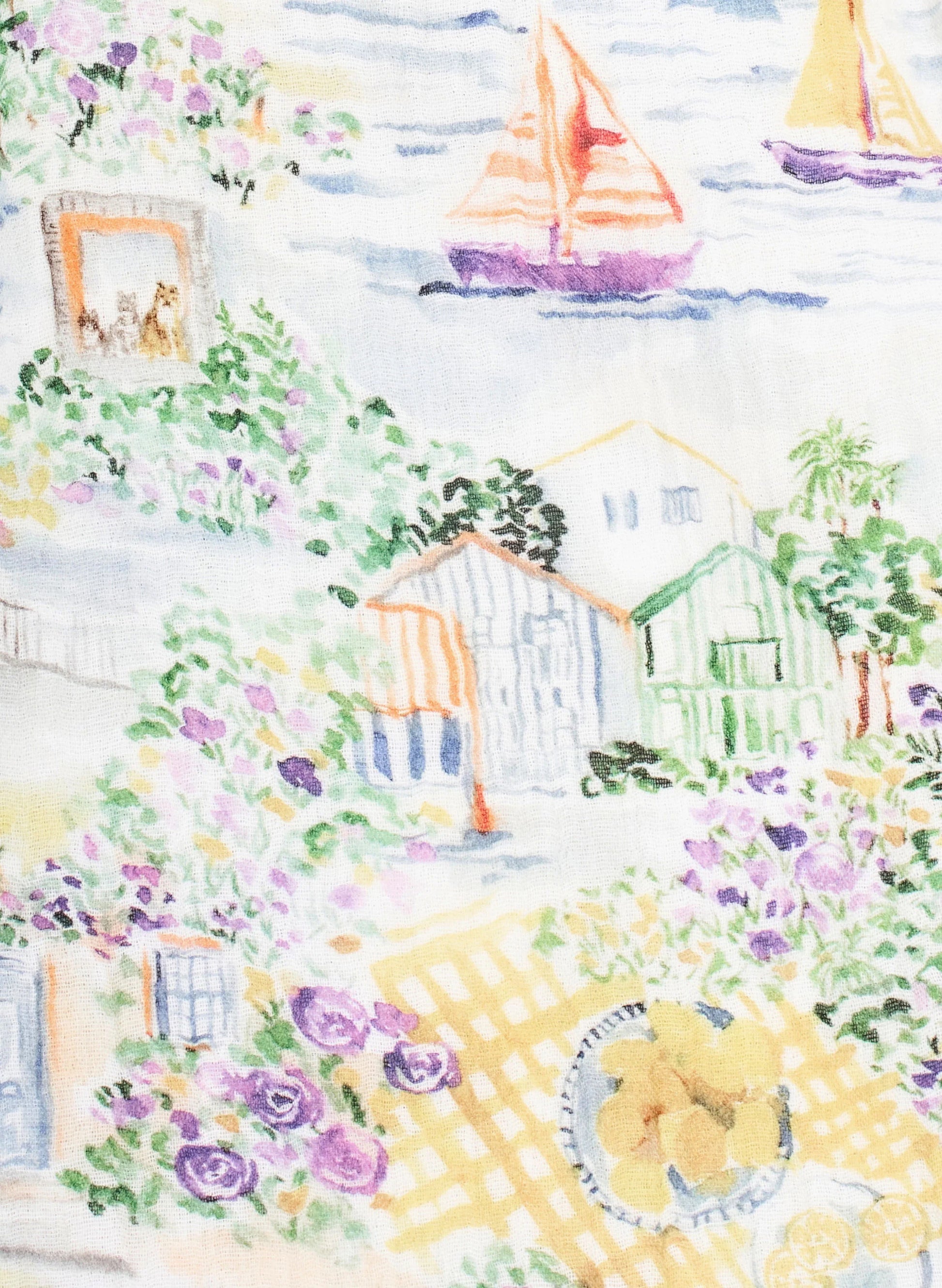 The Timo & Violet Shawl Blanket features a seaside village with colorful houses, sailboats on the water, and flowers and fruit on an organic cotton checkered tablecloth in the foreground.