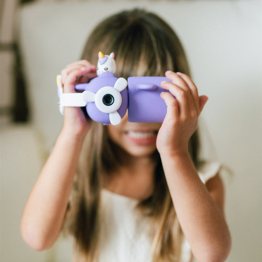 A young child holds a Kidamento Digital Camera Model V, Iris the Unicorn to their eyes, partially obscuring their face. The playful device even has a small unicorn figure on top, adding a touch of magic.