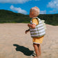 A young child wearing a Sunnylife Kids' Swim Vest—featuring a neoprene comfort guard and anti-rise safety strap—stands on a sandy beach with green hills and a blue sky in the background.