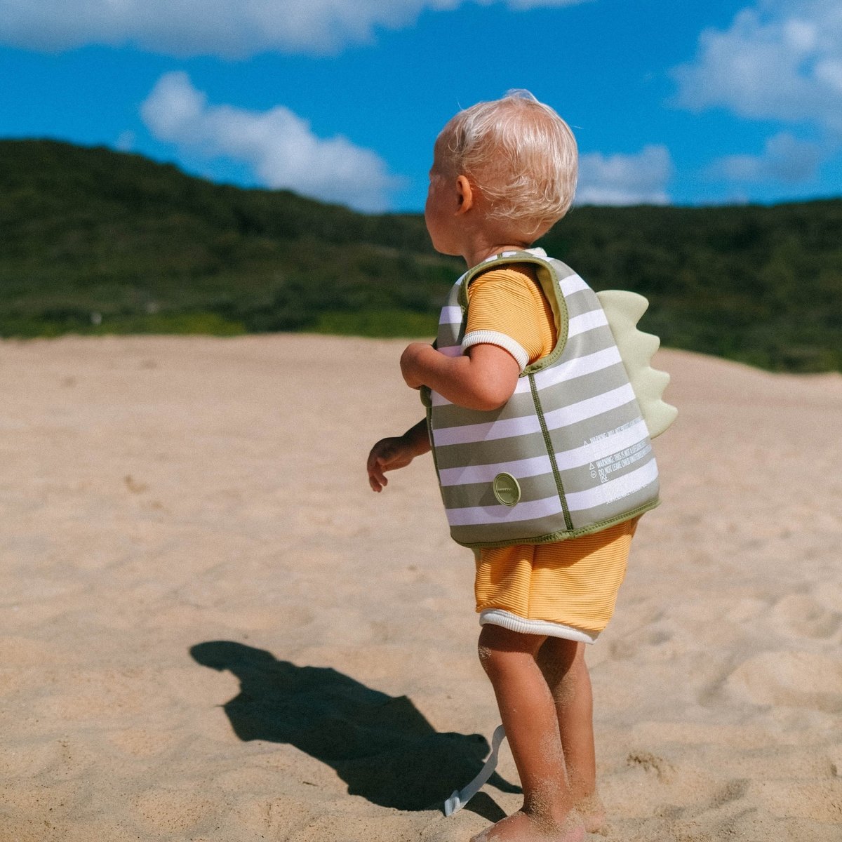 A young child wearing a Sunnylife Kids' Swim Vest—featuring a neoprene comfort guard and anti-rise safety strap—stands on a sandy beach with green hills and a blue sky in the background.