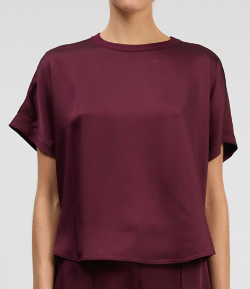 A person wears the Simkhai Addy Satin T-Shirt, a relaxed fit maroon blouse with short sleeves and a round neckline, shown waist up against a plain background.