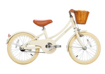 The Banwood Classic Children’s Bicycle is cream-colored with a brown seat, cream tires, and a wicker basket on the front handlebars, displayed in profile against a white background.