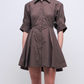 A person stands against a plain background in the Simkhai Jazzie Mini Dress, a brown, short-sleeved, collared button-down with a flared skirt made from soft 100% cotton poplin.
